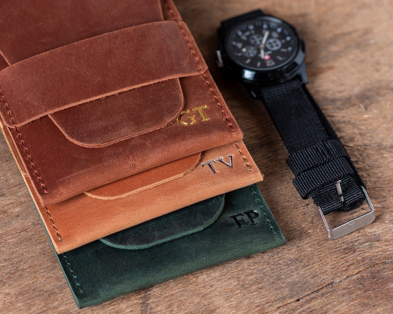 May include: Three leather wallets stacked on top of each other. The top wallet is brown, the middle wallet is tan, and the bottom wallet is green. Each wallet has a different set of initials embossed on the front: GT, TV, and FP.