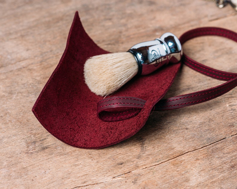 May include: A burgundy leather shaving brush holder with a strap. The holder is empty, but a silver shaving brush with white bristles is lying on top of it.