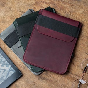 May include: Leather e-reader sleeves in burgundy, gray, and dark green, each with a black elastic band closure. The sleeves are stacked on a wooden surface, with an e-reader and glasses visible in the background. The sleeves are designed to protect e-readers.