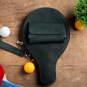 May include: A dark green leather ping pong paddle-shaped bag with a small pocket and a wrist strap. A white ping pong ball and a yellow ping pong ball are next to the bag. A red and blue ping pong paddle is in the lower left corner.