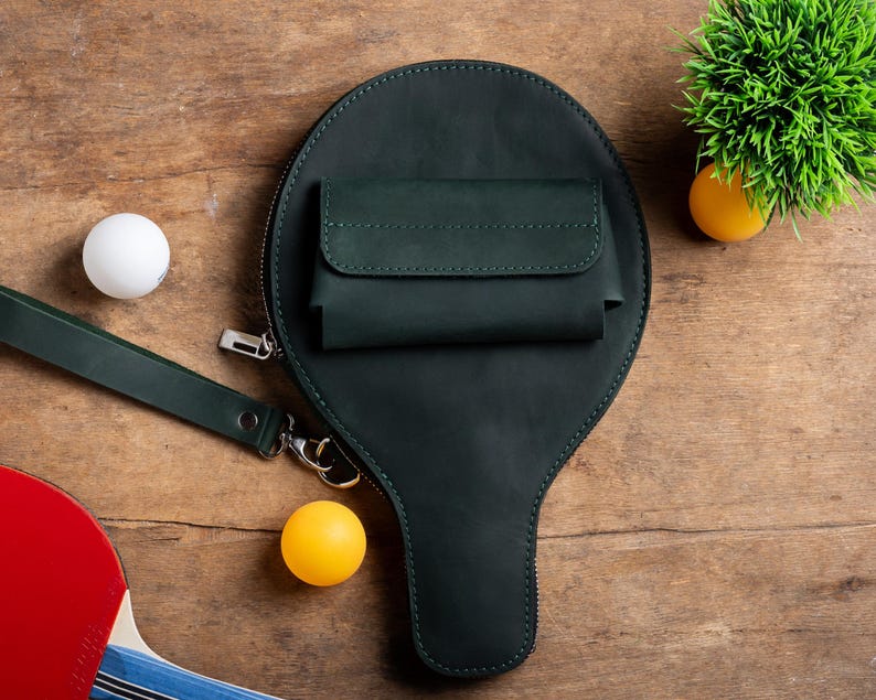 May include: A dark green leather ping pong paddle-shaped bag with a small pocket and a wrist strap. A white ping pong ball and a yellow ping pong ball are next to the bag. A red and blue ping pong paddle is in the lower left corner.