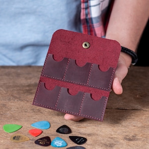 May include: A burgundy leather guitar pick holder with six slots for storing picks. The holder is open and the slots are visible.