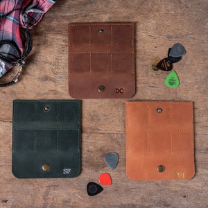 May include: Three leather guitar pick holders with different colored leather. The holders have six slots for picks and a snap closure. The holders are personalized with the initials "DC", "SF", and "VI".