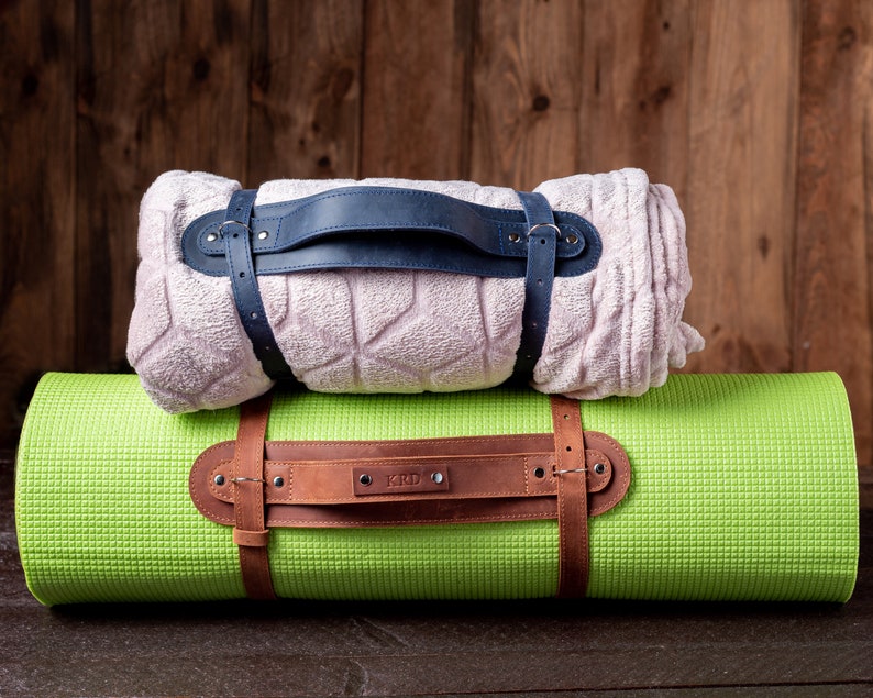 May include: A green yoga mat with a brown leather strap and a blue strap holding a light pink blanket on top.