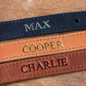 May include: Three leather dog collars with different names embossed on them. The top collar is black with silver lettering that says "MAX". The middle collar is brown with gold lettering that says "COOPER". The bottom collar is dark brown with gold lettering that says "CHARLIE".