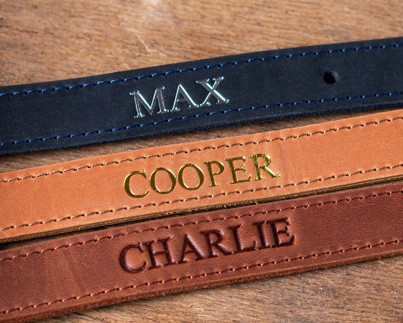 May include: Three leather dog collars with different names embossed on them. The top collar is black with silver lettering that says "MAX". The middle collar is brown with gold lettering that says "COOPER". The bottom collar is dark brown with gold lettering that says "CHARLIE".