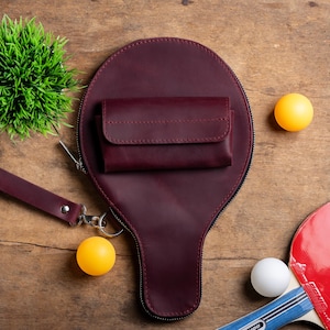 May include: A burgundy leather wallet and paddle-shaped bag with a zipper, a wrist strap, and a small wallet. Two yellow ping pong balls, a white ping pong ball, and a red and black ping pong paddle are also visible.