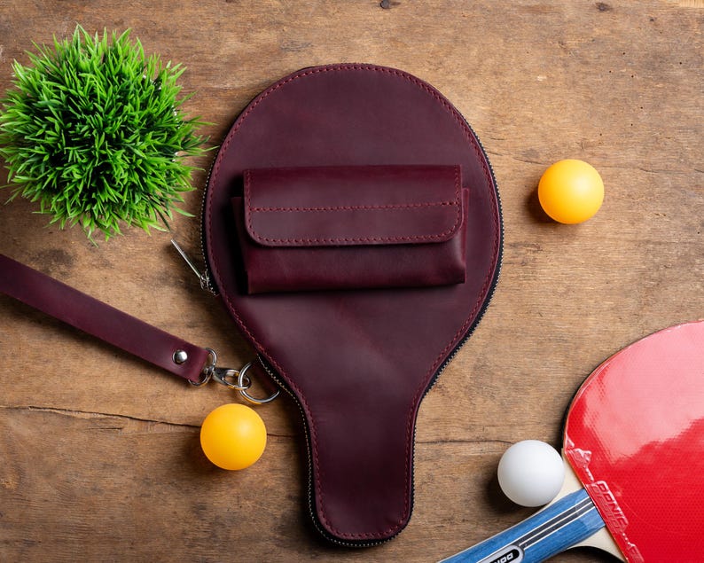 May include: A burgundy leather wallet and paddle-shaped bag with a zipper, a wrist strap, and a small wallet. Two yellow ping pong balls, a white ping pong ball, and a red and black ping pong paddle are also visible.