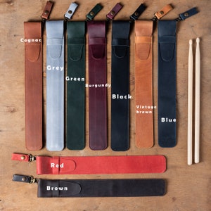 Puede incluir: Surtido de porta baquetas de cuero en varios colores, incluyendo co&ntilde;ac, gris, verde, burdeos, negro, marr&oacute;n vintage y azul. Los porta baquetas est&aacute;n alineados en una superficie de madera con un par de baquetas de madera a la derecha.