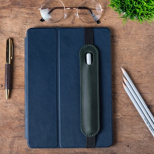 May include: A dark blue leather case with a green leather strap and a pocket for a stylus. The case is on a wooden surface with a pen and pencils nearby.