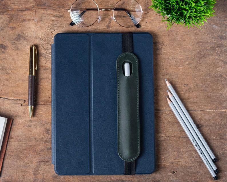 May include: A dark blue leather case with a green leather strap and a pocket for a stylus. The case is on a wooden surface with a pen and pencils nearby.
