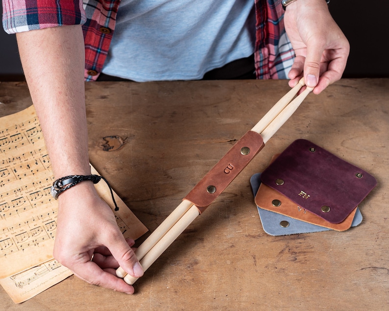May include: A brown leather strap with two wooden drumsticks attached. The strap has the initials "CU" embossed on it. Three leather coasters with initials are in the background.