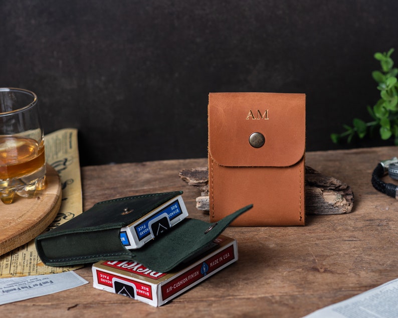 May include: A brown leather card case with the initials "AM" embossed on the front. The case is closed and sitting on a wooden surface next to a deck of playing cards in a green leather case.