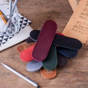 May include: A collection of eight personalised leather pen cases in various colours, including red, brown, green, blue, and grey. Each case has a different monogrammed initial.
