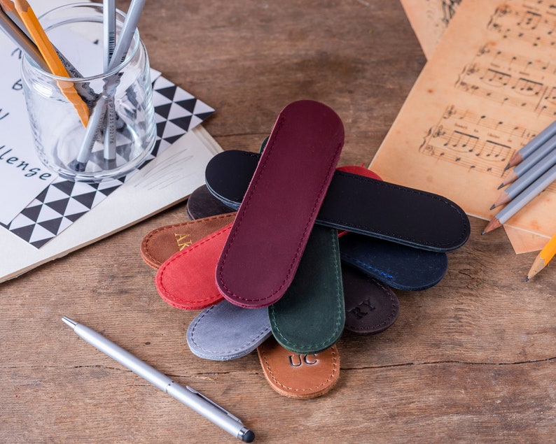 May include: A collection of eight personalised leather pen cases in various colours, including red, brown, green, blue, and grey. Each case has a different monogrammed initial.