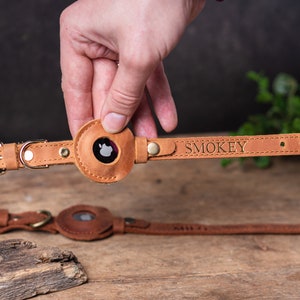 Op de afbeelding: Een bruine leren hondenriem met een ronde, bruine leren AirTag-houder. De riem is gegraveerd met de naam "SMOKEY".