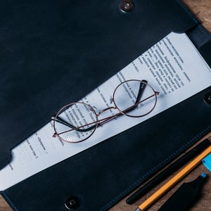 May include: A dark blue leather folder with a white sheet of paper inside. The paper has text written in a foreign language. A pair of round, brown-rimmed glasses are on top of the paper. A black pen, a yellow pencil, and a blue highlighter are on the folder.