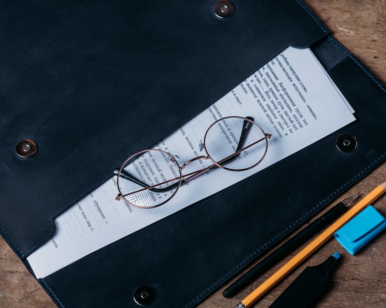May include: A dark blue leather folder with a white sheet of paper inside. The paper has text written in a foreign language. A pair of round, brown-rimmed glasses are on top of the paper. A black pen, a yellow pencil, and a blue highlighter are on the folder.