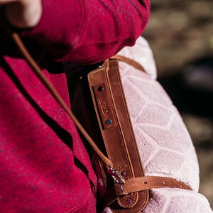 May include: A brown leather strap with a personalised name tag that reads "KRD" is attached to a pink blanket with a geometric pattern. The strap is worn over a red sweater.