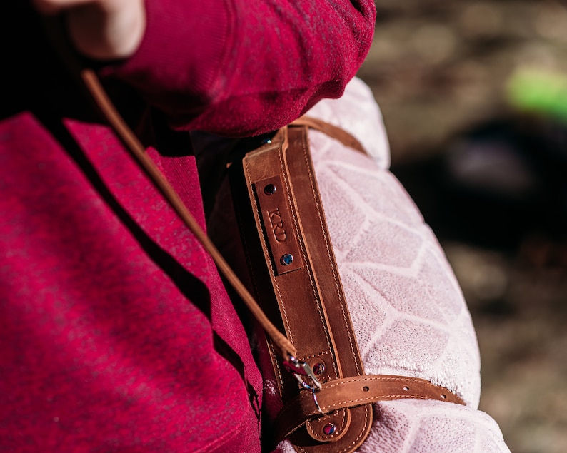 May include: A brown leather strap with a personalised name tag that reads "KRD" is attached to a pink blanket with a geometric pattern. The strap is worn over a red sweater.