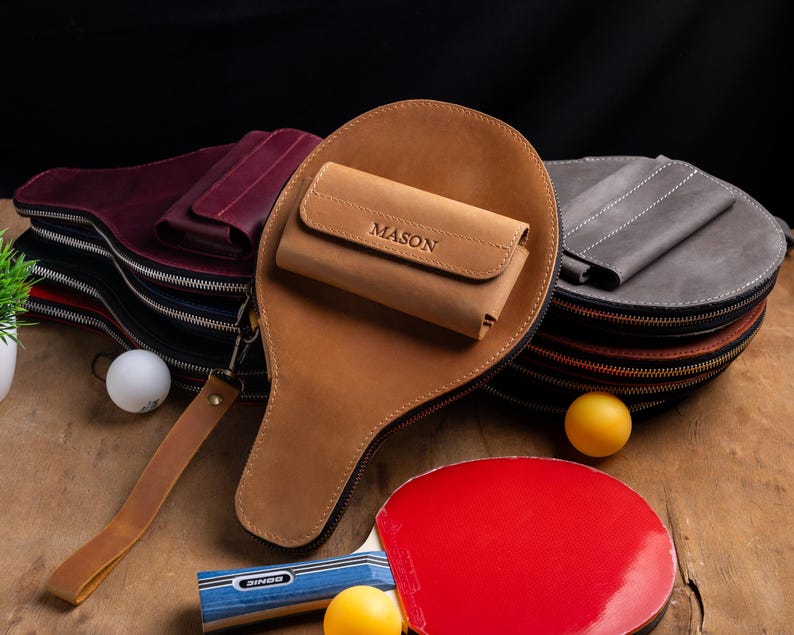 May include: A collection of leather ping pong paddle cases in various colors, including brown, burgundy, and gray. Each case has a zipper closure and a wrist strap. A small leather wallet with the name "MASON" is on top of a brown case. Ping pong balls and paddles are also visible.