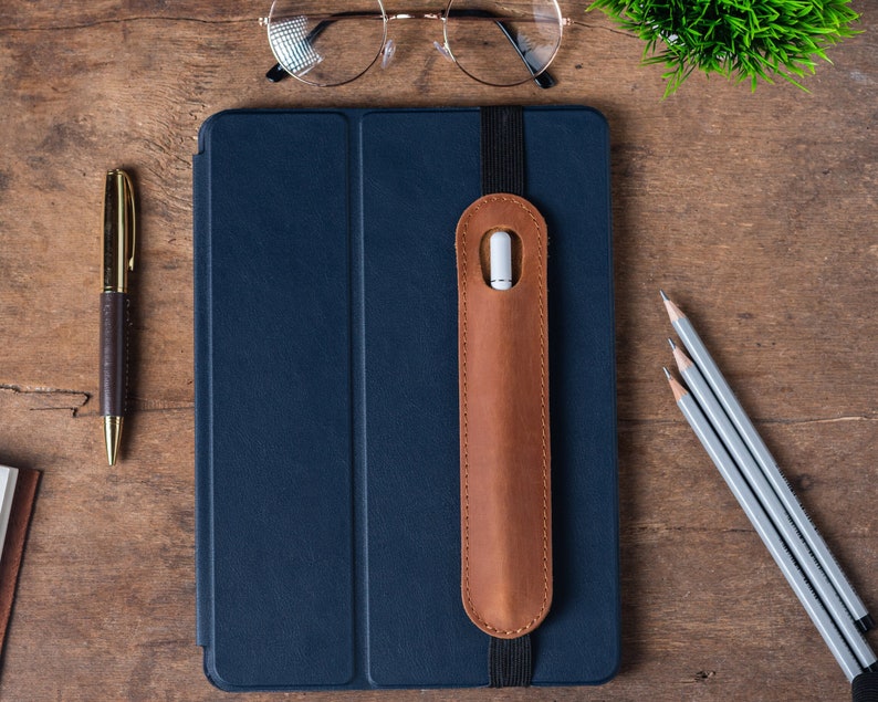 May include: A dark blue tablet case with a brown leather pencil holder attached. The case has a flap that closes with a magnetic closure. The pencil holder has a white pencil inside. There are three pencils and a pen on a wooden surface.