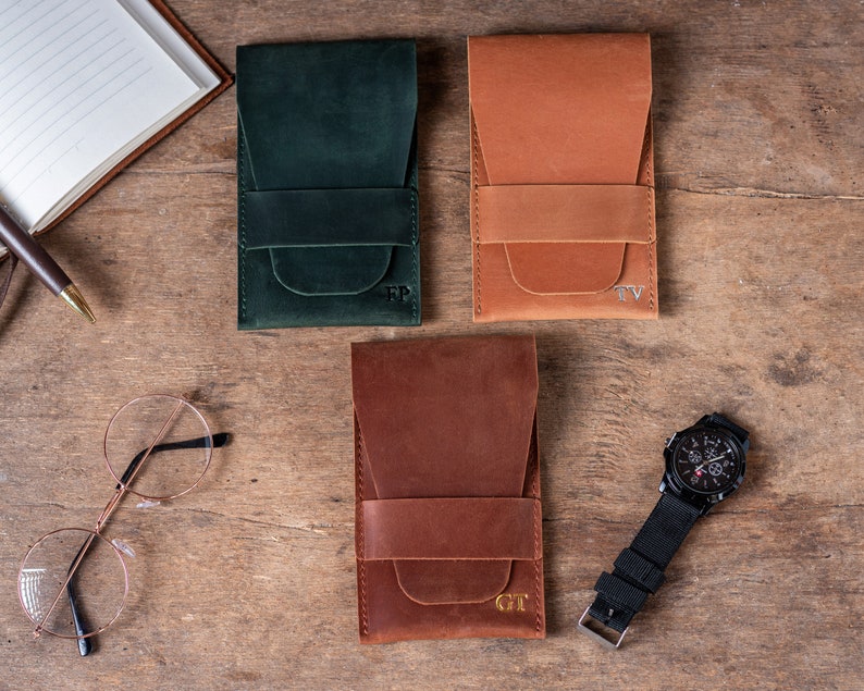 May include: Three leather watch cases in green, brown, and orange with personalized initials. The cases have a flap that closes with a strap. The cases are on a wooden surface.