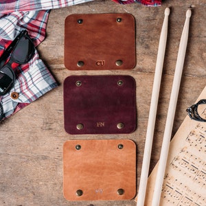 May include: Three leather guitar picks with initials "CU", "FN", and "VY" engraved on them. The picks are brown, burgundy, and tan. The picks are on a wooden surface with a pair of drumsticks and a sheet of music.