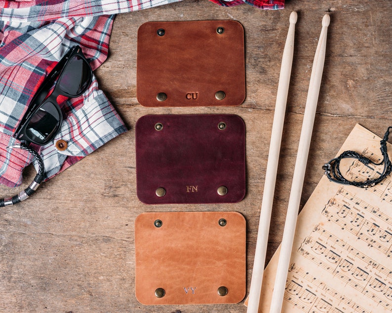 May include: Three leather guitar picks with initials "CU", "FN", and "VY" engraved on them. The picks are brown, burgundy, and tan. The picks are on a wooden surface with a pair of drumsticks and a sheet of music.