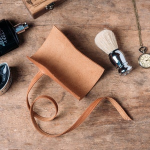 May include: A brown leather shaving brush holder with a strap. The holder is empty and lying on a wooden surface. There is a shaving brush, a bottle of cologne, and a pocket watch on the surface.
