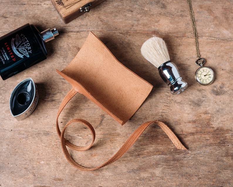 May include: A brown leather shaving brush holder with a strap. The holder is empty and lying on a wooden surface. There is a shaving brush, a bottle of cologne, and a pocket watch on the surface.