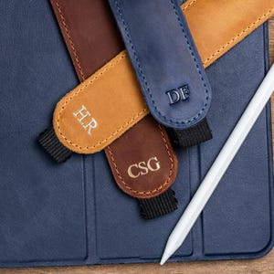 May include: A white stylus rests on a blue tablet case, with three personalized leather pen holders in brown, tan, and blue. Each holder features embroidered initials: HR, DF, and CSG. The holders have black elastic bands.