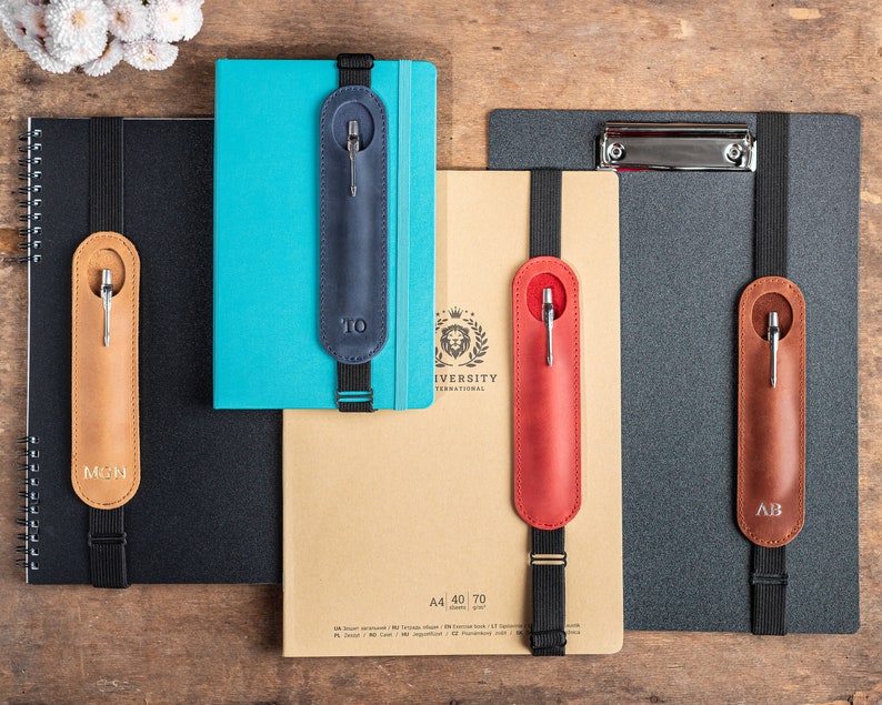 K&ouml;nnte beinhalten: Vier verschiedene Leder-Stift-Halter mit elastischen B&auml;ndern, die an Notizb&uuml;chern und einer Klemmbrett befestigt sind. Die Stift-Halter sind in verschiedenen Farben: braun, rot, blau und beige. Die Stift-Halter haben Initialen: TO, AB, MN und UNIVERSITY INTERNATIONAL.