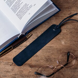 May include: A blue leather bookmark with a black tassel, a gold pen, and a pair of round glasses on a wooden surface.