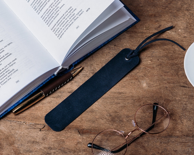 May include: A blue leather bookmark with a black tassel, a gold pen, and a pair of round glasses on a wooden surface.