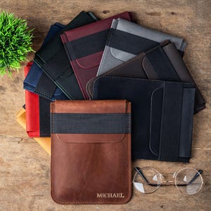 May include: A collection of leather cardholders in various colors, including brown, black, and blue, with elastic bands. The cardholders are arranged on a wooden surface, with a small green plant and a pair of round eyeglasses.