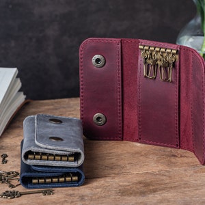 May include: Two leather keychains, one burgundy and one gray, with multiple key rings. The burgundy keychain is open, showing the inside, and the gray keychain is closed. There are several keys scattered on a wooden surface.