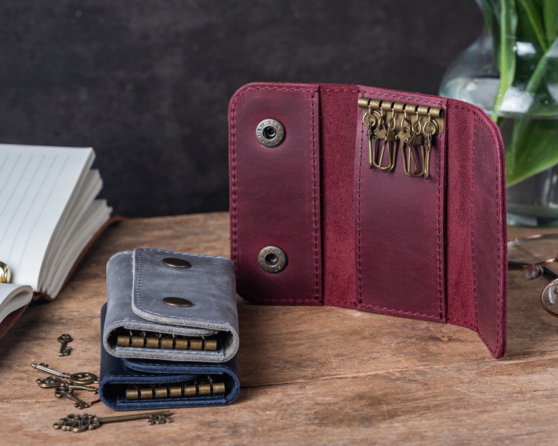 May include: Two leather keychains, one burgundy and one gray, with multiple key rings. The burgundy keychain is open, showing the inside, and the gray keychain is closed. There are several keys scattered on a wooden surface.