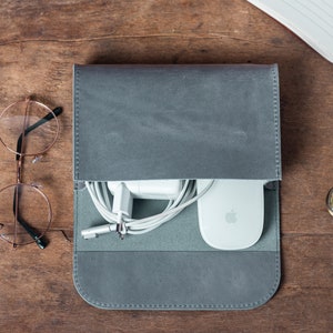 May include: A gray leather tech organizer with a flap, holding a white charger, mouse, and cables. A gold pen, round glasses, and a pocket watch are on a wooden surface.