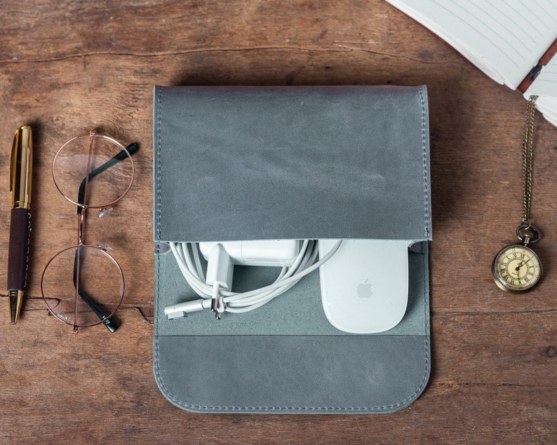 May include: A gray leather tech organizer with a flap, holding a white charger, mouse, and cables. A gold pen, round glasses, and a pocket watch are on a wooden surface.
