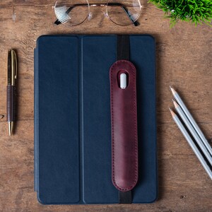 May include: A dark blue leather-bound notebook with a burgundy leather pencil holder attached to the cover. The notebook is on a wooden surface with a gold pen, three pencils, and a pair of glasses.
