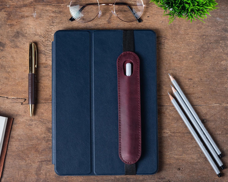 May include: A dark blue leather-bound notebook with a burgundy leather pencil holder attached to the cover. The notebook is on a wooden surface with a gold pen, three pencils, and a pair of glasses.