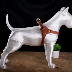 Puede incluir: Escultura de un perro blanco con un arnés de cuero marrón. El perro está sobre una superficie de madera, con una pequeña planta en maceta en el fondo. El arnés tiene un anillo metálico y está asegurado con remaches.