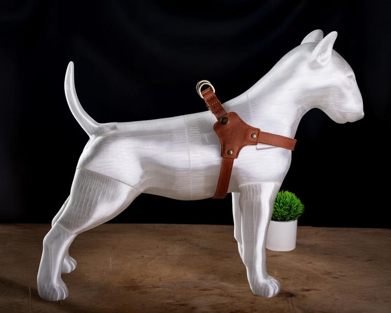 May include: A white dog sculpture with a brown leather harness. The dog is standing on a wooden surface with a small potted plant in the background. The harness has a metal ring and is secured with rivets.