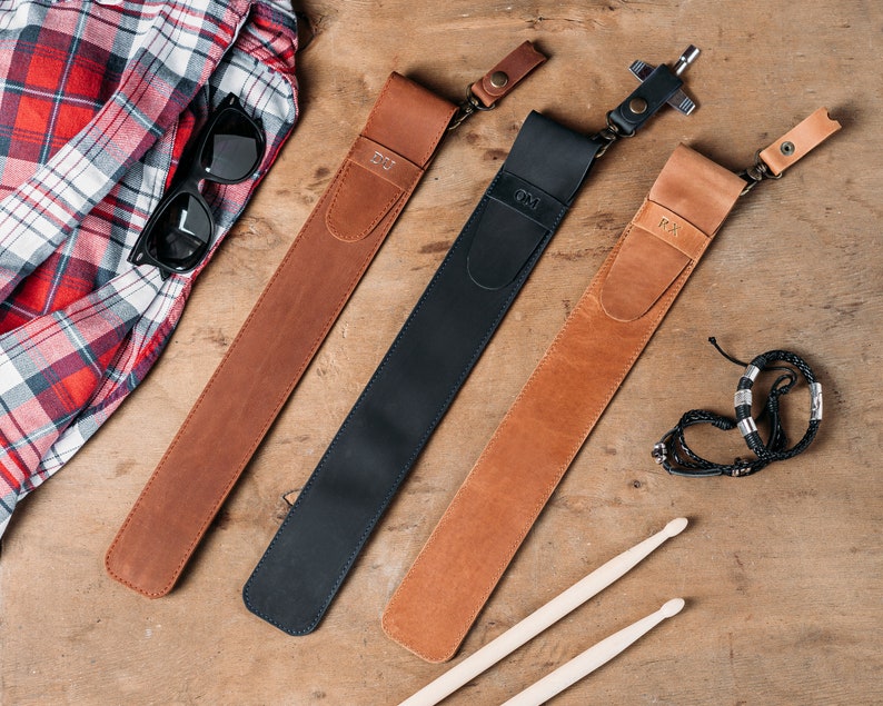 Puede incluir: Tres correas de cuero con iniciales personalizadas, "DJ", "OM" y "PX", est&aacute;n dispuestas sobre una superficie de madera. Las correas son marr&oacute;n, negra y beige. Tambi&eacute;n se puede ver un par de baquetas y un brazalete de cuentas.