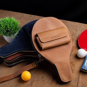 May include: A collection of ping pong accessories, including a brown leather paddle case with a matching wallet, a red and blue ping pong paddle, and a yellow ping pong ball. The wallet is embossed with the name "MASON".