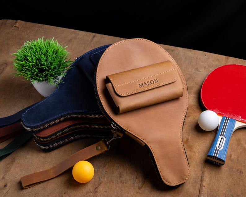 May include: A collection of ping pong accessories, including a brown leather paddle case with a matching wallet, a red and blue ping pong paddle, and a yellow ping pong ball. The wallet is embossed with the name "MASON".