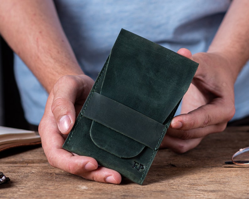 May include: A dark green leather wallet with a flap closure and a single strap. The wallet has the initials "FP" embossed on the front.