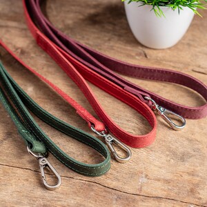 May include: Three leather straps in green, red and burgundy with silver clasps. The straps are lying on a wooden surface.
