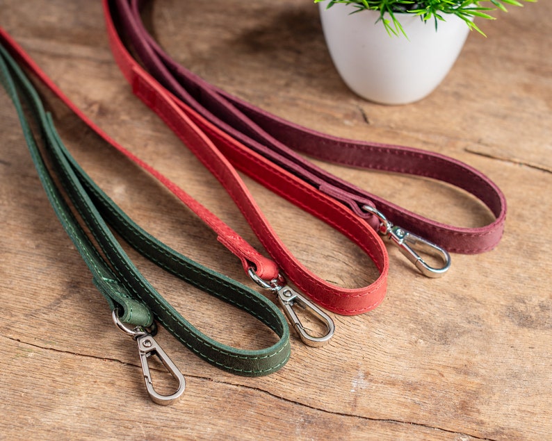 May include: Three leather straps in green, red and burgundy with silver clasps. The straps are lying on a wooden surface.